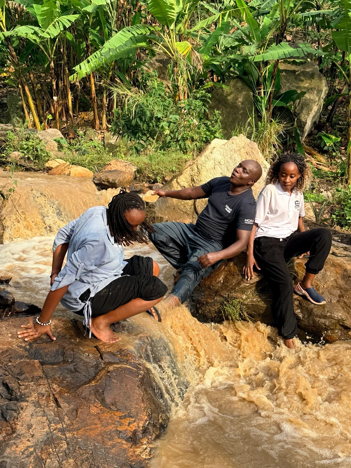 Three people on rock beside a fast-flowing brown stream, with banana plants growing behind them